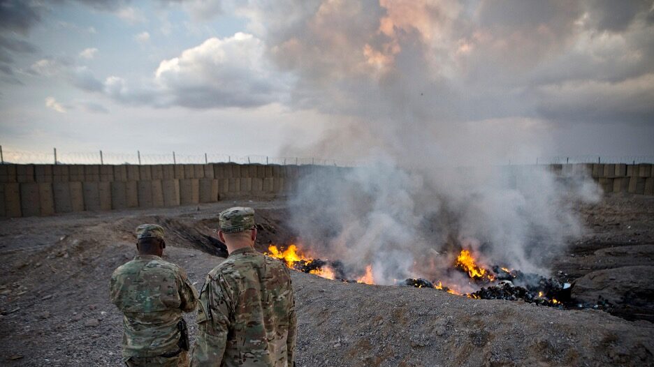 Light grey smoke rises from a giant smouldering pit. Two soliders in fatigues look on. A wall of HESCO barriers and barbed wire encircles one side of the scene.